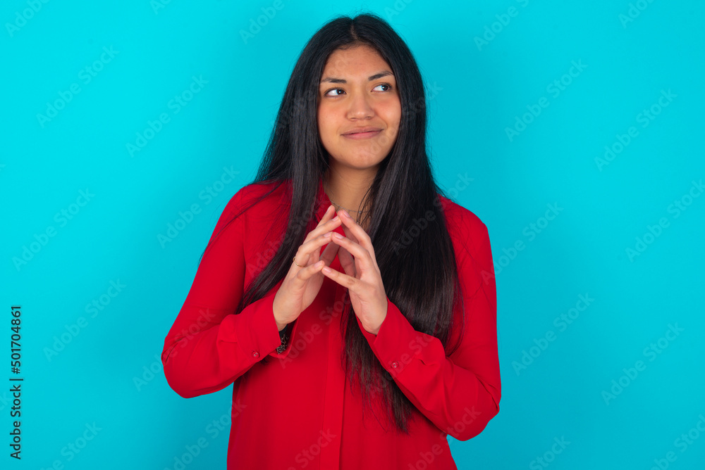 young latin woman wearing red shirt over blue background steepled fingers and looks mysterious aside has great evil plan in mind young latin woman wearing red shirt over blue background steepled fingers and looks mysterious aside has great evil plan in mind