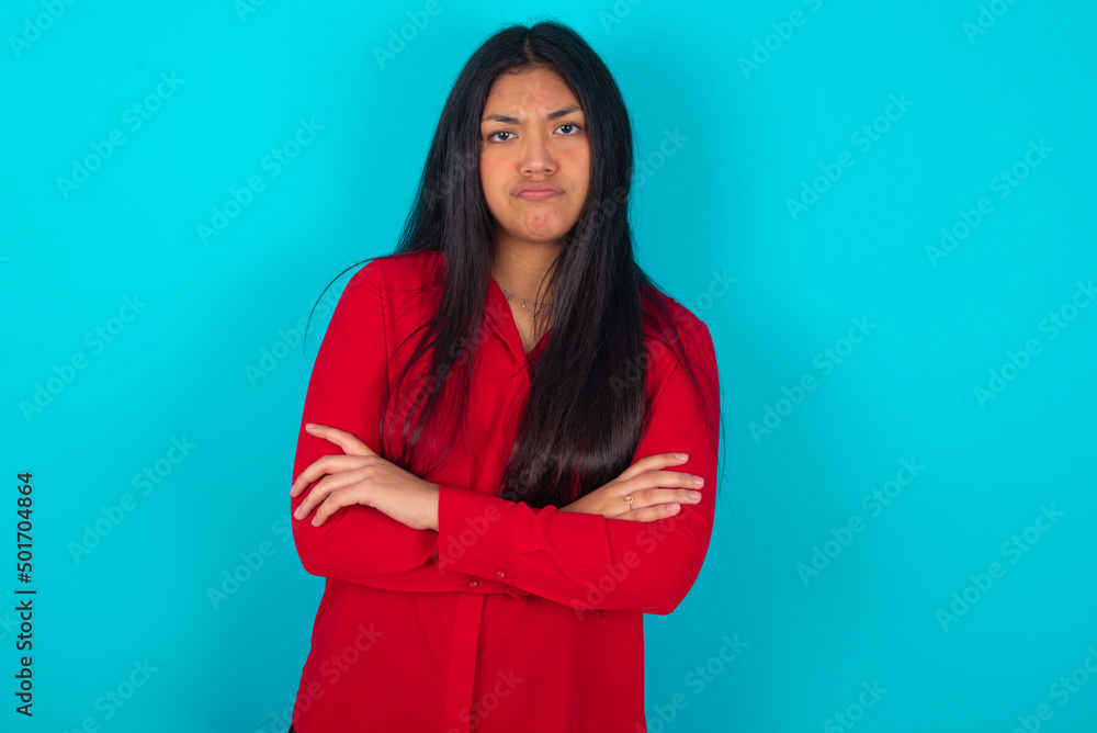 Gloomy dissatisfied young latin woman wearing red shirt over blue background looks with miserable expression at camera from under forehead, makes unhappy grimace Gloomy dissatisfied young latin woman wearing red shirt over blue background looks with miserable expression at camera from under forehead, makes unhappy grimace