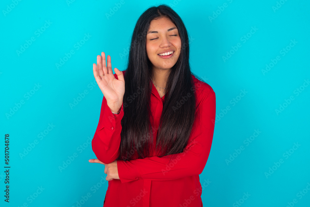 Fototapeta premium Overjoyed successful young latin woman wearing red shirt over blue background raises palm and closes eyes in joy being entertained by friends