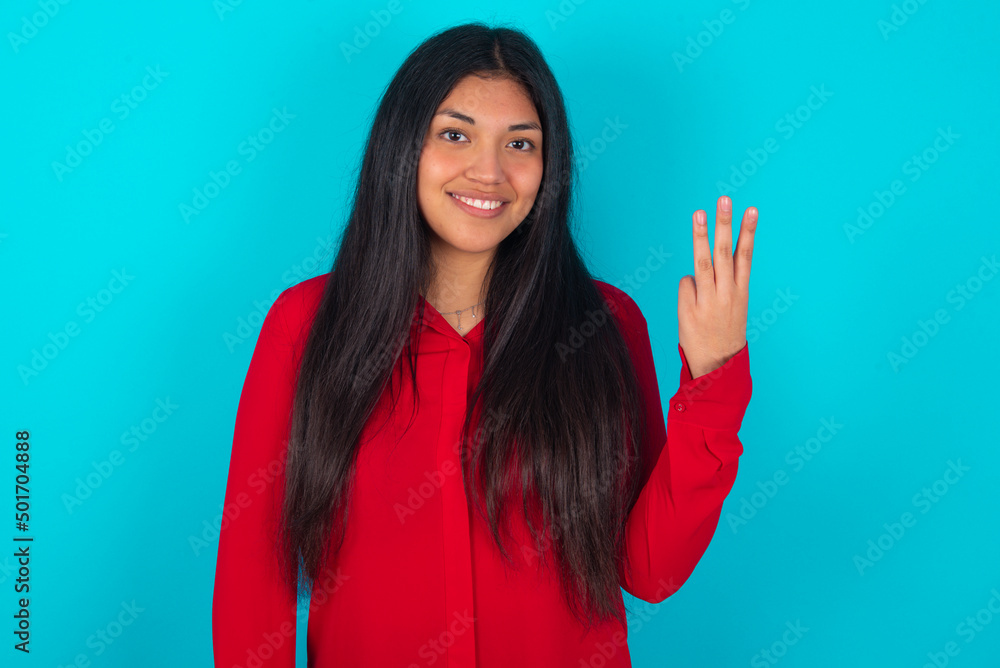 young latin woman wearing red shirt over blue background smiling and looking friendly, showing number three or third with hand forward, counting down young latin woman wearing red shirt over blue background smiling and looking friendly, showing number three or third with hand forward, counting down