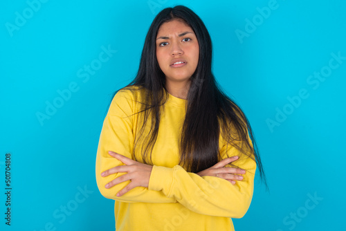 young latin woman wearing yellow sweater over blue background frowning his face in displeasure, keeping arms folded, waiting for an explanation.