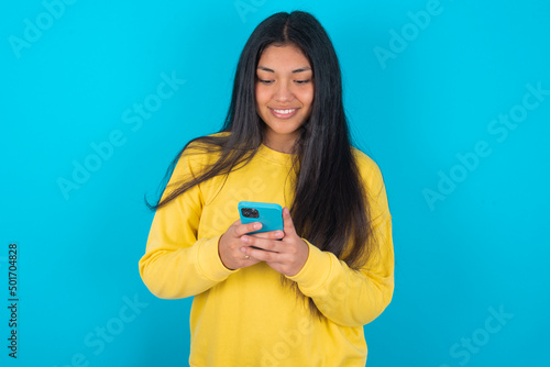 Smiling young latin woman wearing yellow sweater over blue background using cell phone, messaging, being happy to text with friends, looking at smartphone. Modern technologies and communication. photo