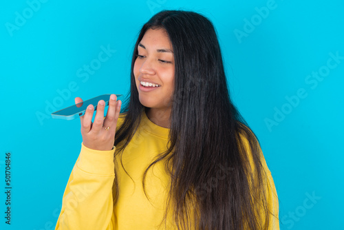 Wallpaper Mural Smiling young latin woman wearing yellow sweater over blue background sending voice message on her smart phone. Communication and new technologies concept. Torontodigital.ca