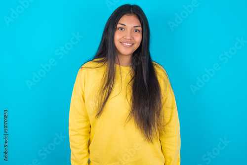 young latin woman wearing yellow sweater over blue background with nice beaming smile pleased expression. Positive emotions concept
