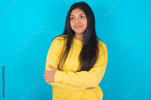Pleased young latin woman wearing yellow sweater over blue background keeps hands crossed over chest looks happily aside