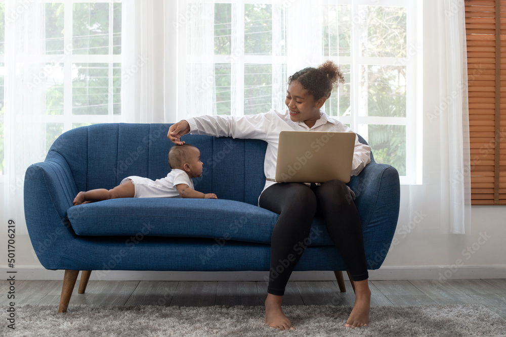 African mother working remotely on laptop while taking care of her baby on couch, freelance single mom working at home, 5 months baby old lying on couch near her African mother working remotely on laptop while taking care of her baby on couch, freelance single mom working at home, 5 months baby old lying on couch near her