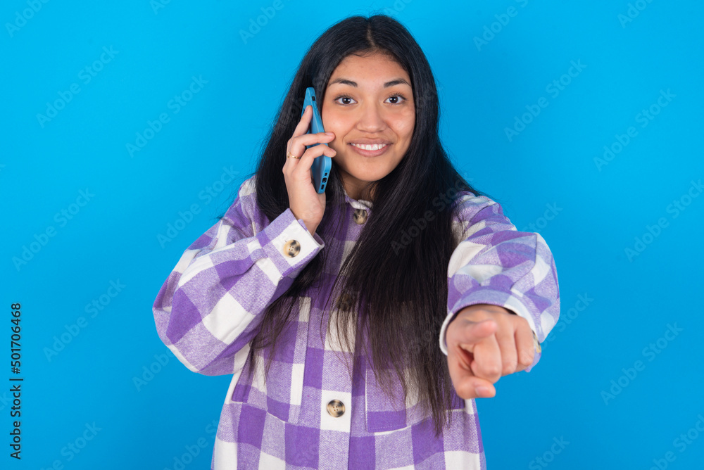 Positive young latin woman wearing plaid shirt over blue background indicates directly at camera has telephone conversation smiles broadly enjoys talking long hours. You join me Positive young latin woman wearing plaid shirt over blue background indicates directly at camera has telephone conversation smiles broadly enjoys talking long hours. You join me