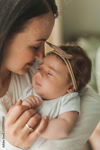 Young beautiful dark-haired mother holds her newborn daughter in her arms in a cozy home. Family portrait. Motherhood. Young woman kisses her 2 month old baby.