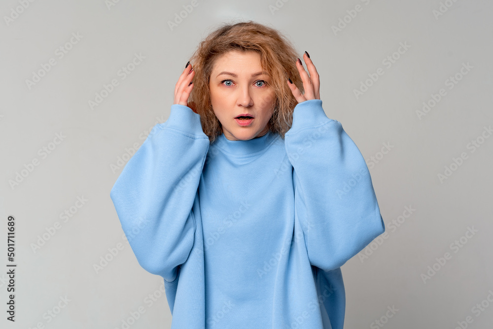 Portrait of scared mature woman looking puzzled and surprised with open mouth as she is shouting while hands are holding head, standing in trendy blue sweatshirt over light grey background Portrait of scared mature woman looking puzzled and surprised with open mouth as she is shouting while hands are holding head, standing in trendy blue sweatshirt over light grey background