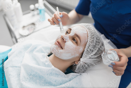 cosmetologist applies a moisturizing white cream mask on her face to a smiling patient with a brush