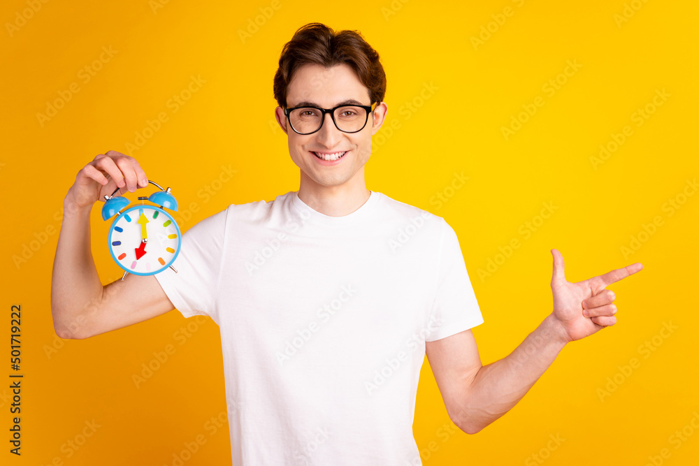 Photo of young guy indicate finger empty space advertise choose hold clock isolated over yellow color background Photo of young guy indicate finger empty space advertise choose hold clock isolated over yellow color background