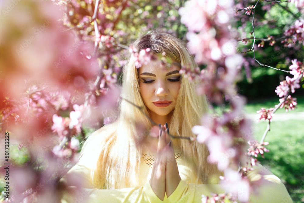 Young blonde woman does yoga outdoors in nature. Beautiful girl makes namaste. Flowering trees in spring. Close-up face with healthy skin among the branches of a sakura tree with flowers in the park. Young blonde woman does yoga outdoors in nature. Beautiful girl makes namaste. Flowering trees in spring. Close-up face with healthy skin among the branches of a sakura tree with flowers in the park.