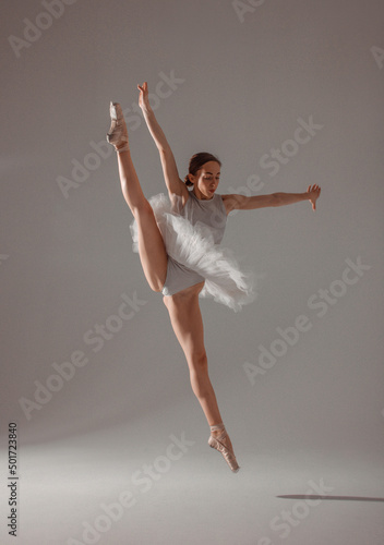 ballet flexible trick. young slim dancer, fit girl ballerina jumps in white ballet tutu like white swan in cross twine in air near the white wall background in studio light. ballet concept, free space