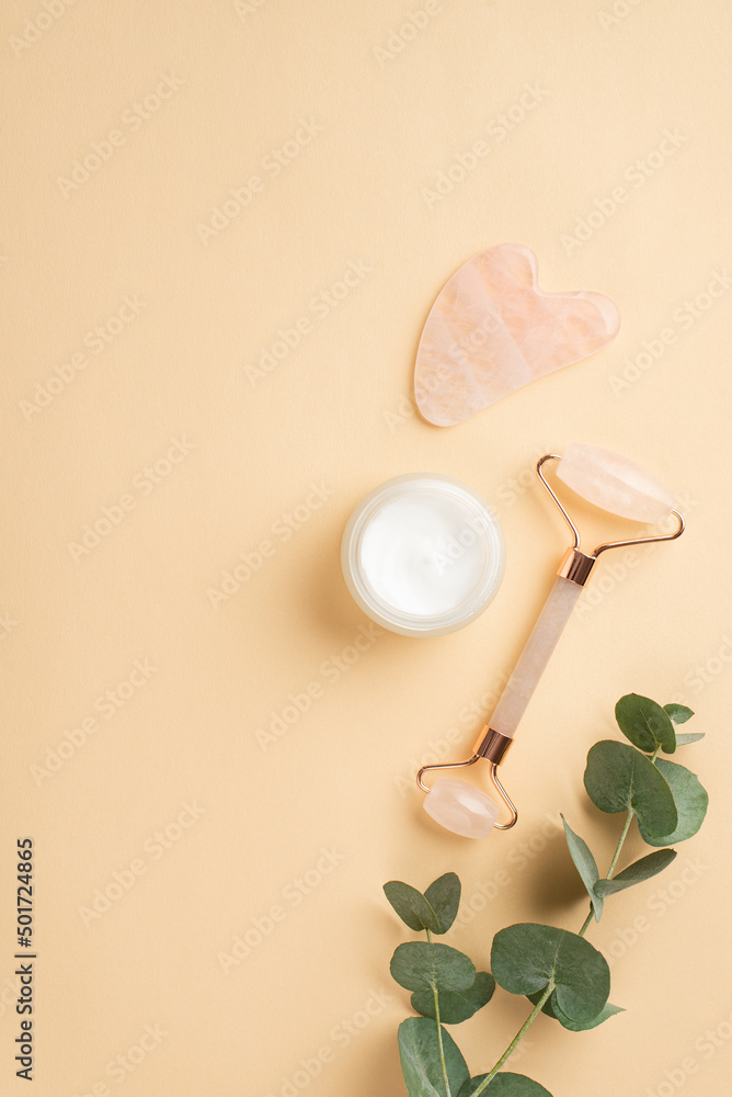 Beauty procedure concept. Top view vertical photo of rose quartz roller gua sha massager glass cream jar and eucalyptus branch on isolated pastel beige background Beauty procedure concept. Top view vertical photo of rose quartz roller gua sha massager glass cream jar and eucalyptus branch on isolated pastel beige background