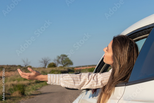 Excited young beautiful woman looking out the car window feeling the air with her arms raised in the countryside