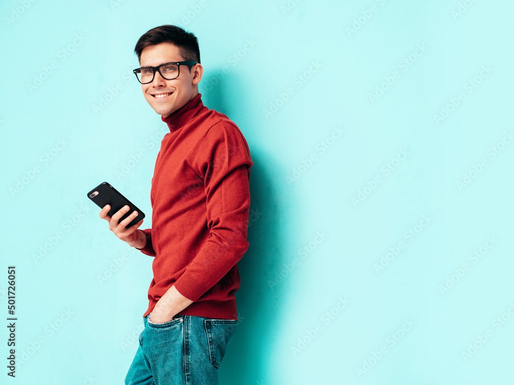 Handsome smiling model. Sexy stylish man dressed in red turtleneck sweater and jeans. Fashion hipster male posing near blue wall in studio.Holding smartphone. Looking at cellphone screen. Using apps Handsome smiling model. Sexy stylish man dressed in red turtleneck sweater and jeans. Fashion hipster male posing near blue wall in studio.Holding smartphone. Looking at cellphone screen. Using apps