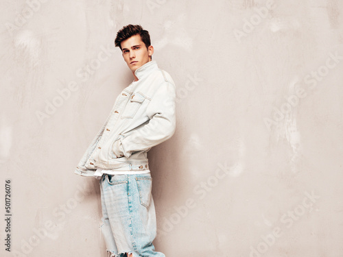 Portrait of handsome confident stylish hipster lambersexual model.Man dressed in jacket and jeans. Fashion male posing in studio near grey wall