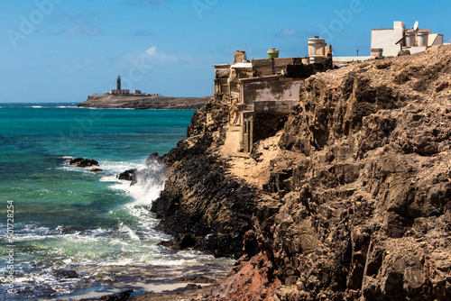 Vista de casas antiguas de Puerto de la Cruz que cuelgan en las rocas y el faro de Punta Jandia en el fondo, Fuerteventura, Islas Canarias, España