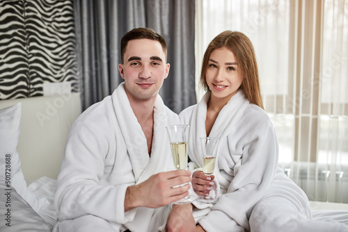 Beauty, spa, healthy lifestyle concept. Beautiful young couple in bathrobes relaxing at luxury hotel room. The boy and the girl are resting in a cozy place and drinking wine. Wellness theme © bondvit