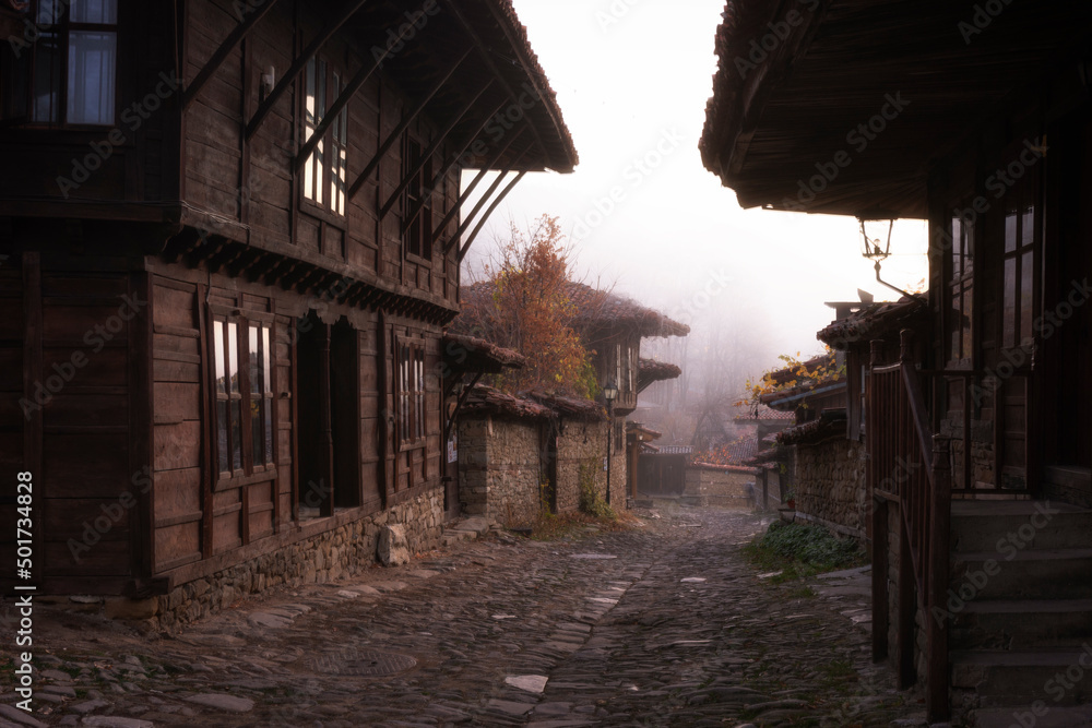 Foggy autumn morning at a street of small village Zheravna in Bulgaria. Traditional historical wooden houses and first light of sun reflecting in the windows. Tourism, travel concept.