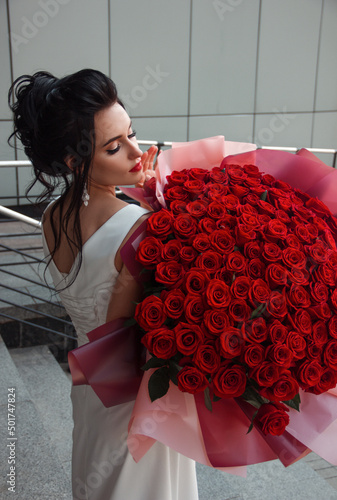 girl celebration. close up young brunette bride in white wedding dress stands casual with huge luxury bouquet of red roses in hands on a gray street building background. lifestyle concept, free space