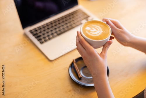 cropped view of young freelancer holding cup of cappuccino near laptop in cafe.