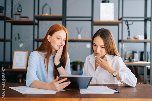 Portrait of two shocked young business women working using digital tablet at meeting desk with job documents at office. Business female colleagues using touchscreen computer for project discussion