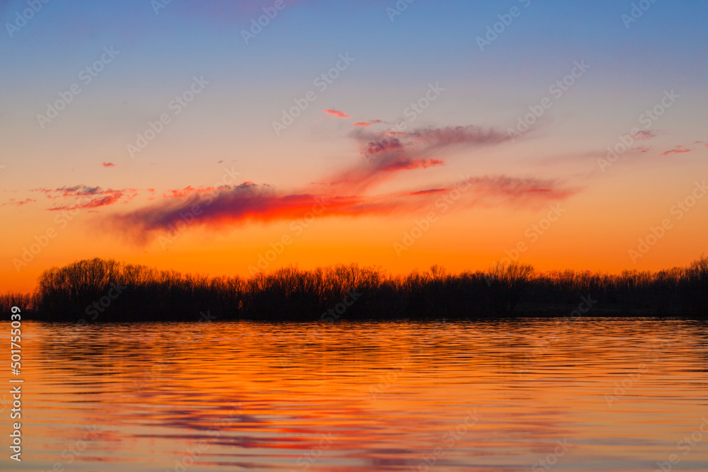 Orange purple and violet sunset on river with dark colorful clouds in sky with trees reflection in water 