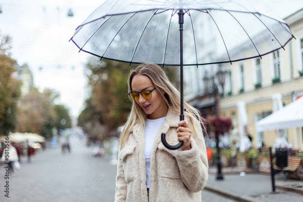 Cute fashion blonde woman in yellow sunglasses with a transparent umbrella walks along the city street. Lifestyle