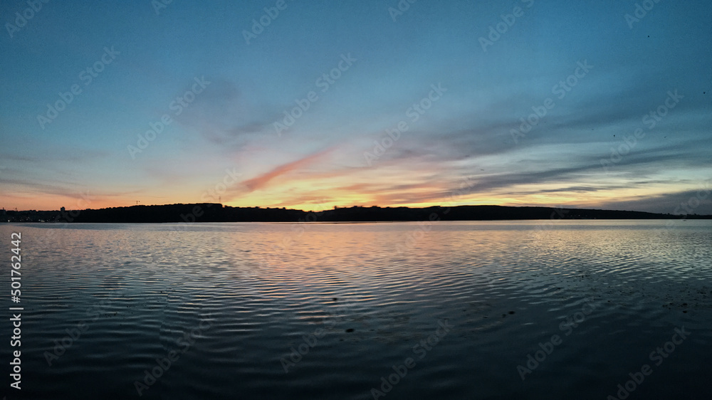 Beautiful sunset over lake Natural background with blue water and sky with yellow orange purple clouds, reflected in the lake. Breathtaking horizon panoramic view with setting sun. Beautiful sunset over lake Natural background with blue water and sky with yellow orange purple clouds, reflected in the lake. Breathtaking horizon panoramic view with setting sun.