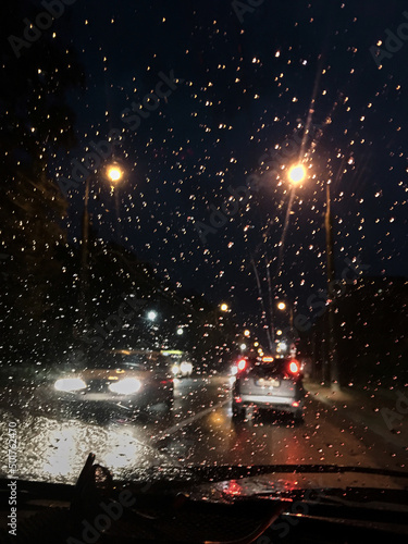 Night city view from the windscreen of the car at night. Rain drops on automobile window. Street lights reflections during summer shower. traveling by car in the evening in city town.