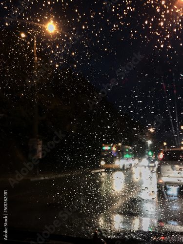 Night city view from the windscreen of the car at night. Rain drops on automobile window. Street lights reflections during summer shower. traveling by car in the evening in city town.