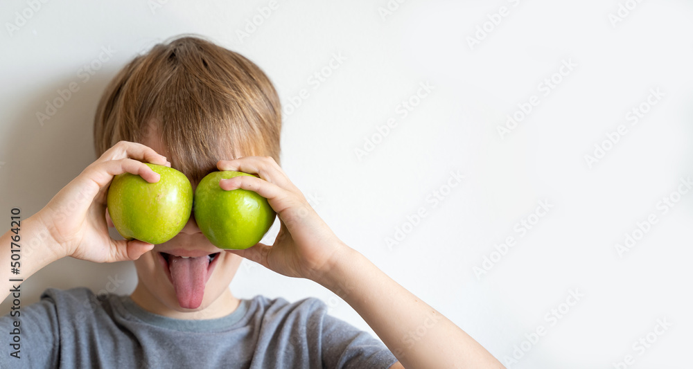 Cute child in grey t-shirt showing tongue and holding green apples in front of his eyes over white background. Concept of healthy nutrition is easy. Cute child in grey t-shirt showing tongue and holding green apples in front of his eyes over white background. Concept of healthy nutrition is easy.