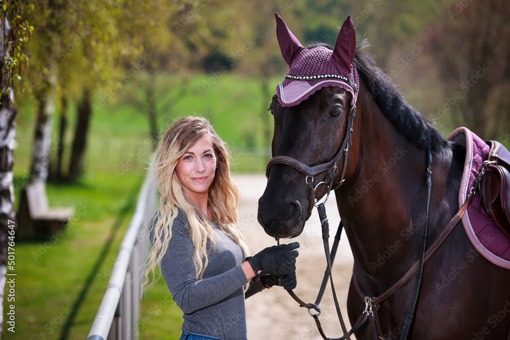 Obraz premium Young woman with long blond hair stands at the edge of a riding arena and holds her horse by the bridle and both look into the camera..