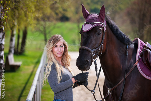Young woman with long blond hair stands at the edge of a riding arena and holds her horse by the bridle and both look into the camera..