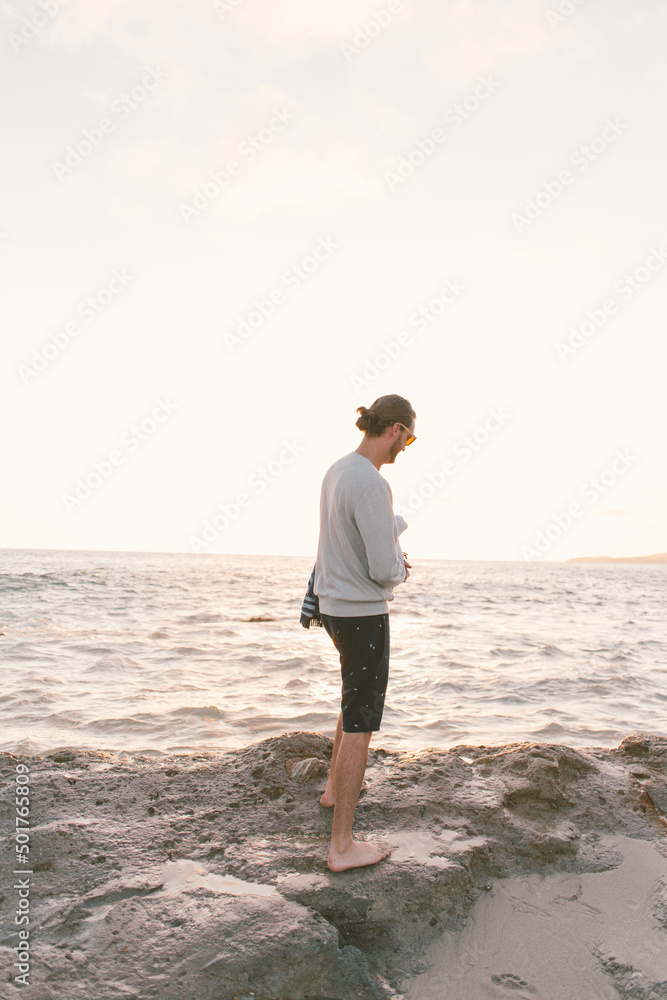 man standing on rocks by ocean water with sunset behind him