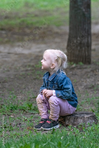 Little blonde girl sits on stump in the woods. Portrait of cute fair-haired girl in the park. Vertical frame.