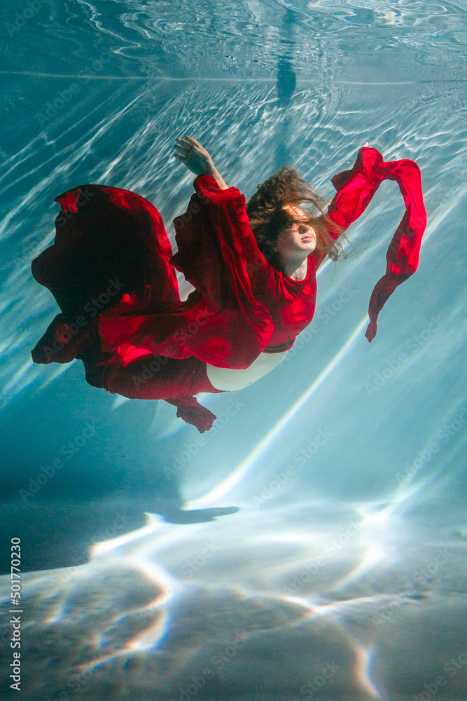 Attractive red-haired young woman swims beautifully underwater in a red ...