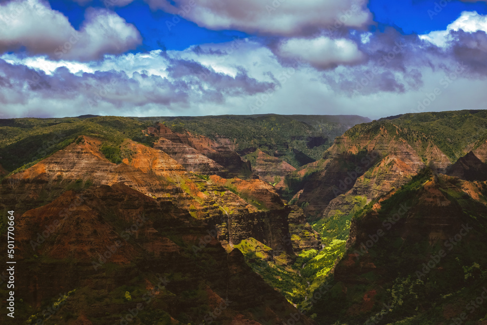 Waimea Canyon on the Hawaiian Island of Kauai, with red rocks and bright green foliage, dark gloomy clouds, bright blue sun, and long beautiful waterfalls 