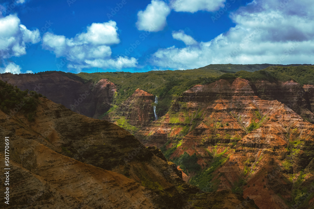 Waimea Canyon on the Hawaiian Island of Kauai, with red rocks and bright green foliage, dark gloomy clouds, bright blue sun, and long beautiful waterfalls 