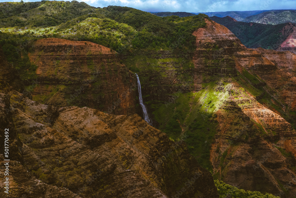 Waimea Canyon on the Hawaiian Island of Kauai, with red rocks and bright green foliage, dark gloomy clouds, bright blue sun, and long beautiful waterfalls 