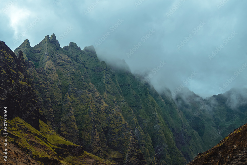The gorgeous rugged wilderness and cliffs of Kauai's Napali Coast in Hawaii, with low clouds and mist hanging over the mountain peaks under a stormy grey sky The gorgeous rugged wilderness and cliffs of Kauai's Napali Coast in Hawaii, with low clouds and mist hanging over the mountain peaks under a stormy grey sky