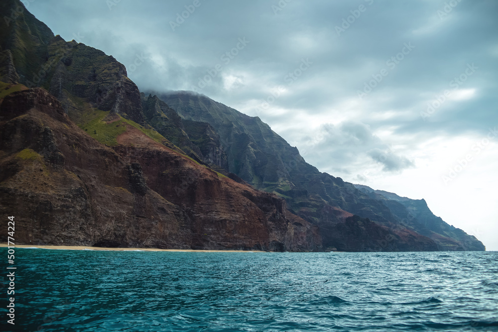 The gorgeous rugged wilderness and cliffs of Kauai's Napali Coast in Hawaii, with low clouds and mist hanging over the mountain peaks under a stormy grey sky, and bright blue and teal ocean waves