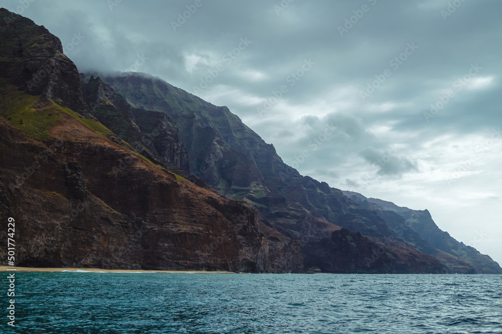 The gorgeous rugged wilderness and cliffs of Kauai's Napali Coast in Hawaii, with low clouds and mist hanging over the mountain peaks under a stormy grey sky, and bright blue and teal ocean waves