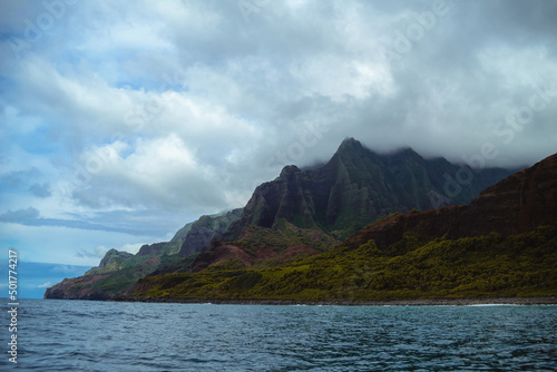 The gorgeous rugged wilderness and cliffs of Kauai's Napali Coast in Hawaii, with low clouds and mist hanging over the mountain peaks under a stormy grey sky, and bright blue and teal ocean waves