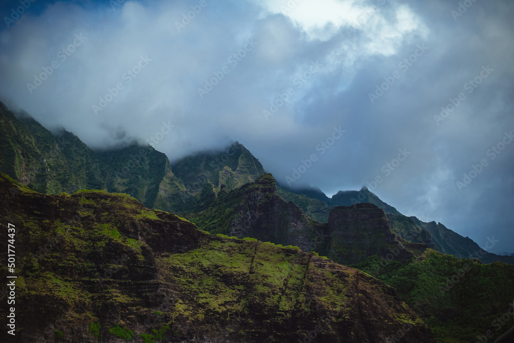 The gorgeous rugged wilderness and cliffs of Kauai's Napali Coast in Hawaii, with low clouds and mist hanging over the mountain peaks under a stormy grey sky The gorgeous rugged wilderness and cliffs of Kauai's Napali Coast in Hawaii, with low clouds and mist hanging over the mountain peaks under a stormy grey sky