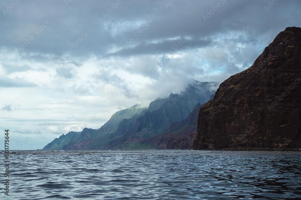 Naklejka premium The gorgeous rugged wilderness and cliffs of Kauai's Napali Coast in Hawaii, with low clouds and mist hanging over the mountain peaks under a stormy grey sky, and bright blue and teal ocean waves