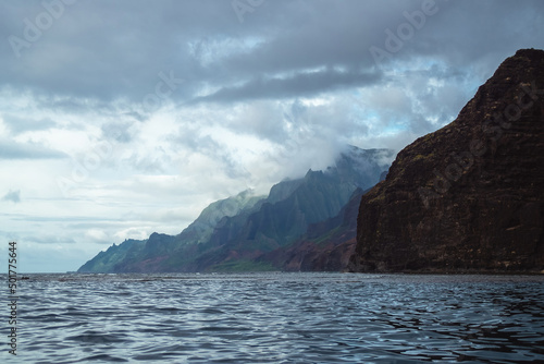 The gorgeous rugged wilderness and cliffs of Kauai's Napali Coast in Hawaii, with low clouds and mist hanging over the mountain peaks under a stormy grey sky, and bright blue and teal ocean waves