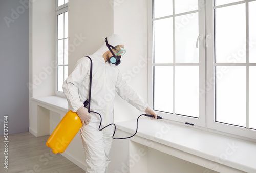 Pest control worker inside the house. Exterminator wearing white protective suit, mask and goggles holding yellow sprayer bottle and spraying insecticide over window sill in modern living room at home