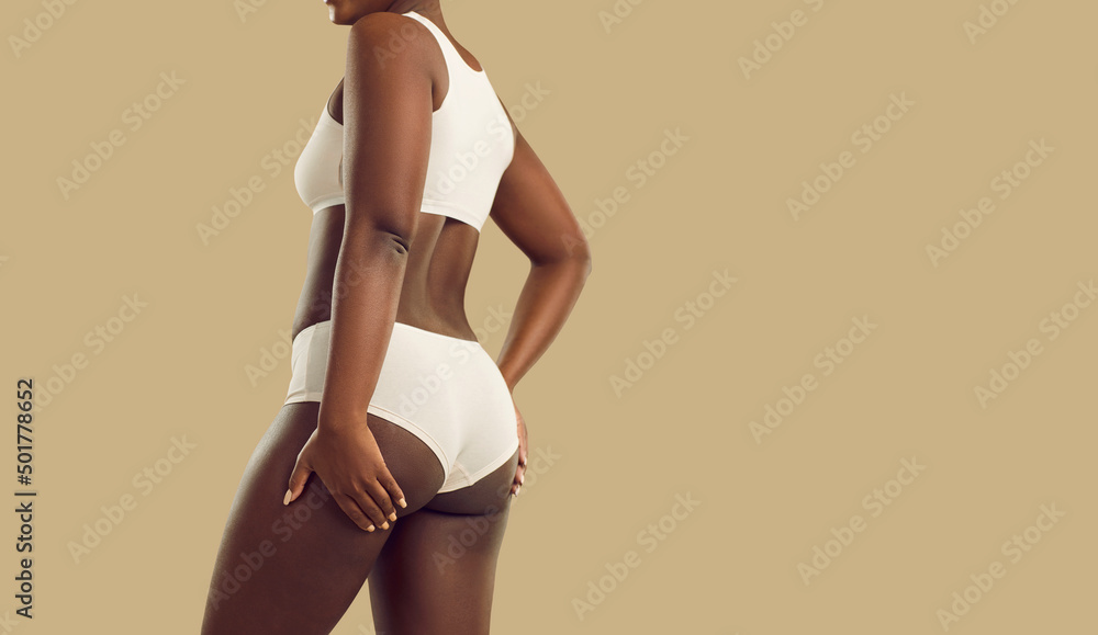 Attractive plus size black woman in underwear. Hot brown skinned model  wearing comfy natural cotton undies holding hands on thighs standing on  color background. Female body concept, rear back view Stock Photo